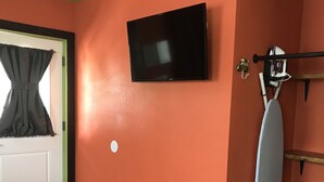 Ein Zimmer mit oranger Wand, einem flachbildfernseher über einer Tür, einem gestreiften Bügeleisen auf einem Gestell und einem Bett mit blumigem Überwurf.
