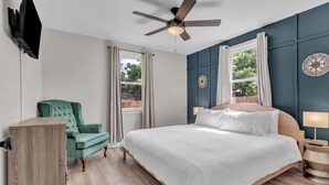Une chambre à coucher comprenant un lit, un bureau, une chaise, un ventilateur de plafond et une fenêtre avec des rideaux.
