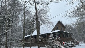 Un chalet rustique doté d’une terrasse en bois et d’un escalier menant à l’entrée, le tout entouré d’arbres couverts de neige.