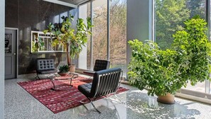 Un interior moderno con una mesa de vidrio, dos sillas negras, un tapete rojo estampado y grandes ventanales con vistas a árboles.