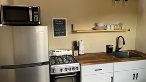 A compact kitchen with a stainless steel refrigerator, a microwave, a gas stove, and a sink. There is a chalkboard with kitchen courtesy rules, a wooden shelf with dishes, and a roll of paper towels.