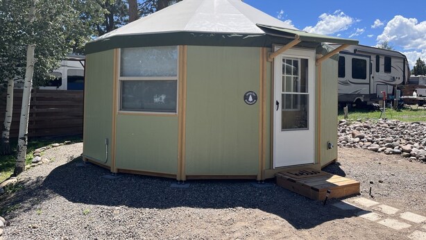 A small, single-story, green-roofed, octagonal structure with a door and windows, situated on gravel with trees and a camper in the background.