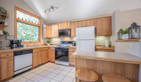 A modern kitchen with wooden cabinets, a dishwasher, an oven, a microwave, and a refrigerator. There is a window providing natural light and a small seating area with stools.