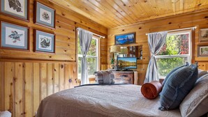 Interior de una cabaña de madera con una cama, cuadros enmarcados, un televisor y una ventana con cortinas.