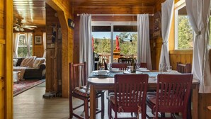 Interior de una cabaña de madera con comedor, sofá y vistas al exterior.