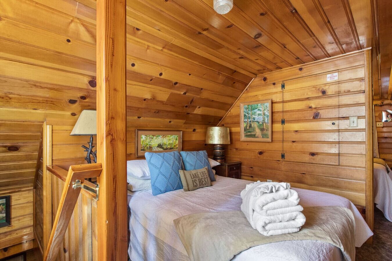 A wooden cabin bedroom with a bed, pillows, a lamp, and a painting on the wall.
