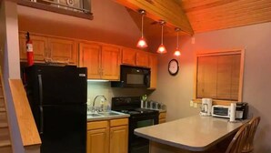 Une cuisine avec des meubles de rangement en bois, un réfrigérateur noir, un four à micro-ondes, une cuisinière et un évier. On y trouve également une poubelle et une horloge au mur.