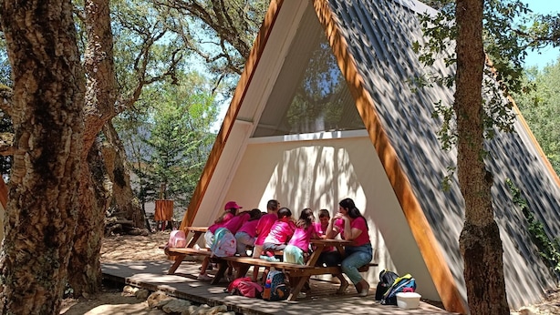 A group of people seated at a wooden table under a triangular tent in a wooded area. - Natura Fun House & Camping (Sintra)