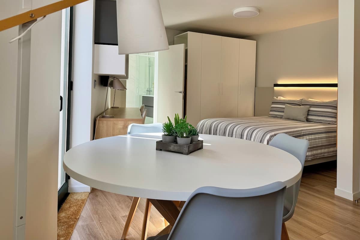 A modern, compact living space with a round dining table, a bed with striped bedding, and a kitchenette in the background.