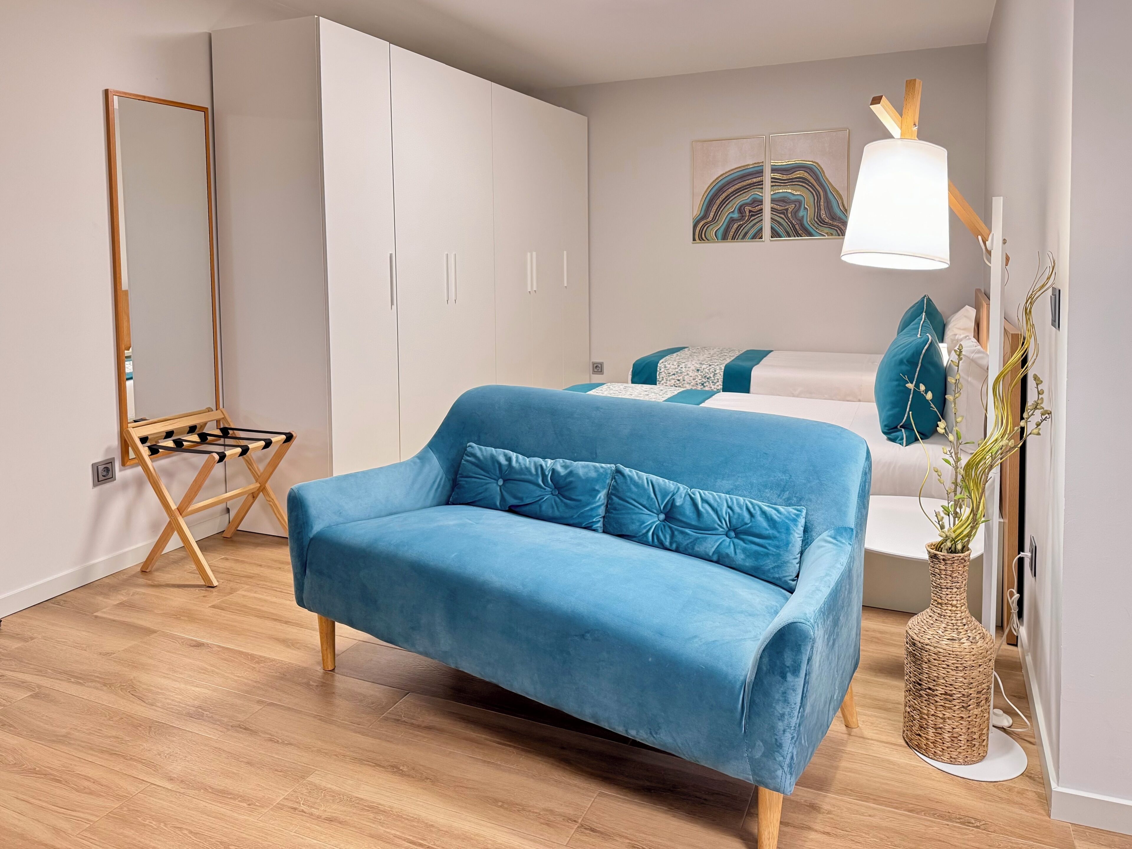 A modern bedroom with a blue sofa, a bed with teal pillows, a large mirror, and a wall-mounted lamp.