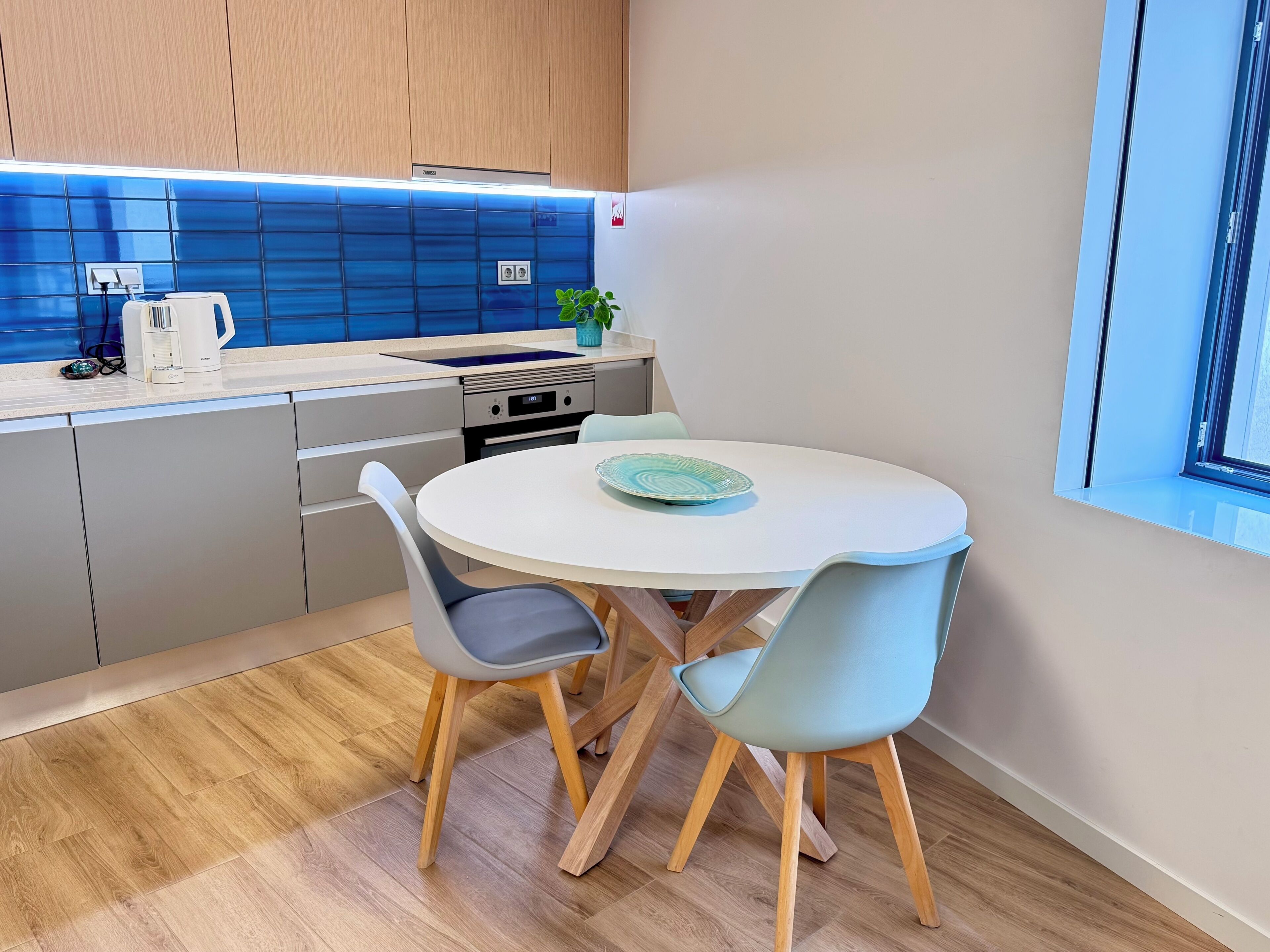 A modern kitchen with a round dining table, two chairs, a coffee maker, and a potted plant.