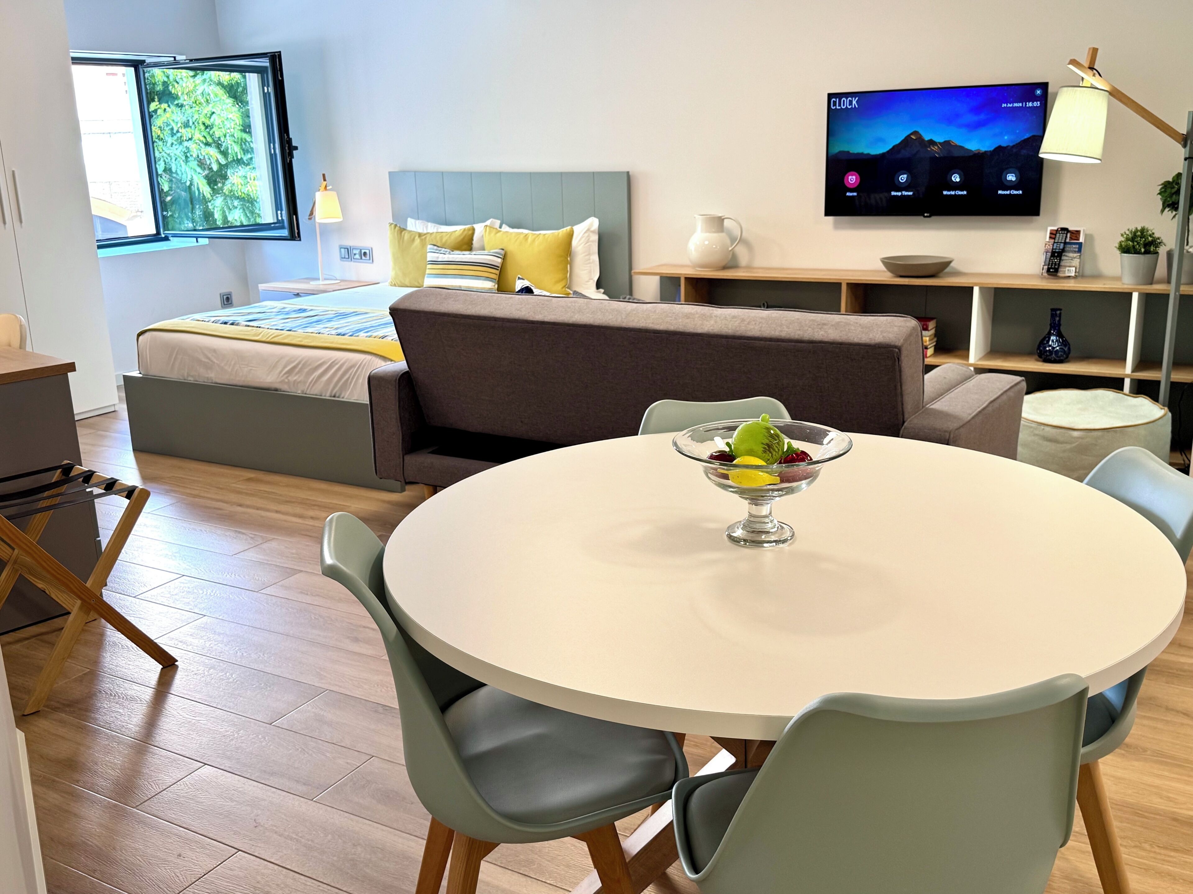 Habitación de hotel moderna con cama, mesa de comedor con sillas, televisor en la pared y ventana con vistas a árboles.