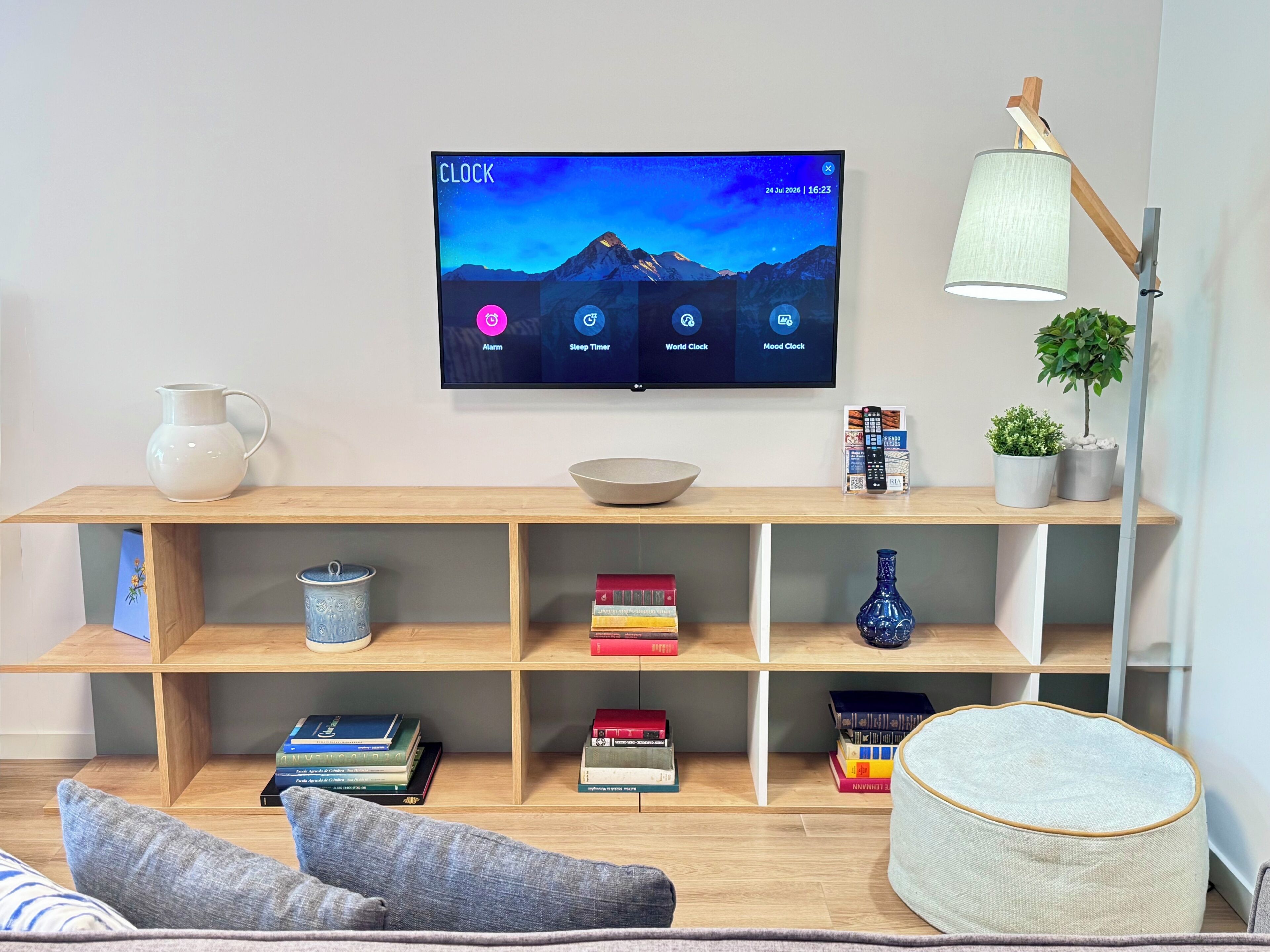 Una sala de estar moderna con un televisor montado mostrando una aplicación de reloj, un estante de madera con libros y adornos, una lámpara de pie y una pequeña planta en maceta.