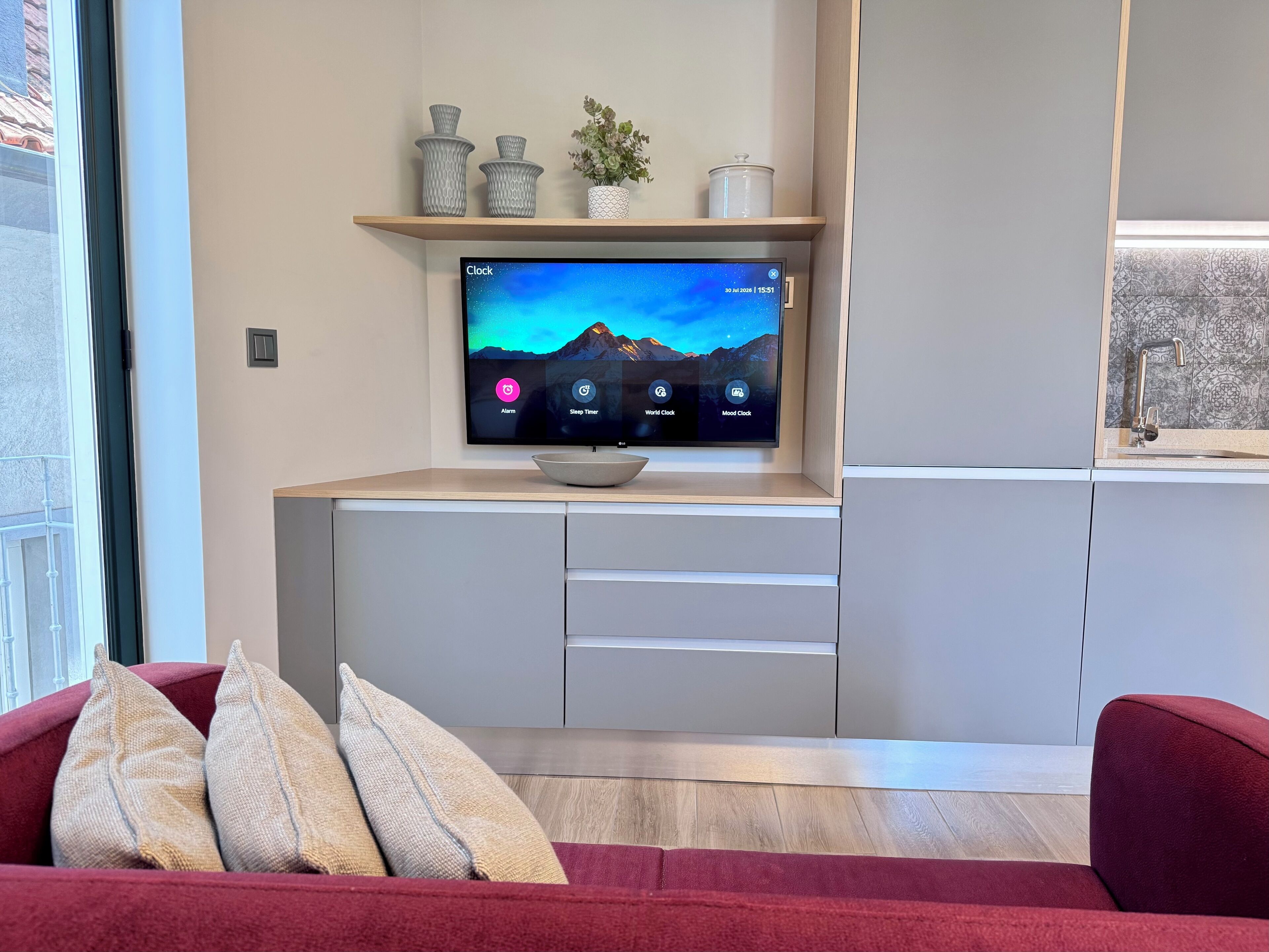 A modern living room with a flat-screen TV mounted on a wooden shelf, a red sofa with cushions, and a kitchen area with a sink and countertop.