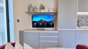 A modern living room with a flat-screen TV mounted on a wooden shelf, a red sofa with cushions, and a kitchen area with a sink and countertop.