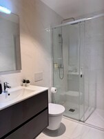 Un baño moderno con ducha de vidrio, un lavamanos blanco con mueble oscuro y un inodoro.