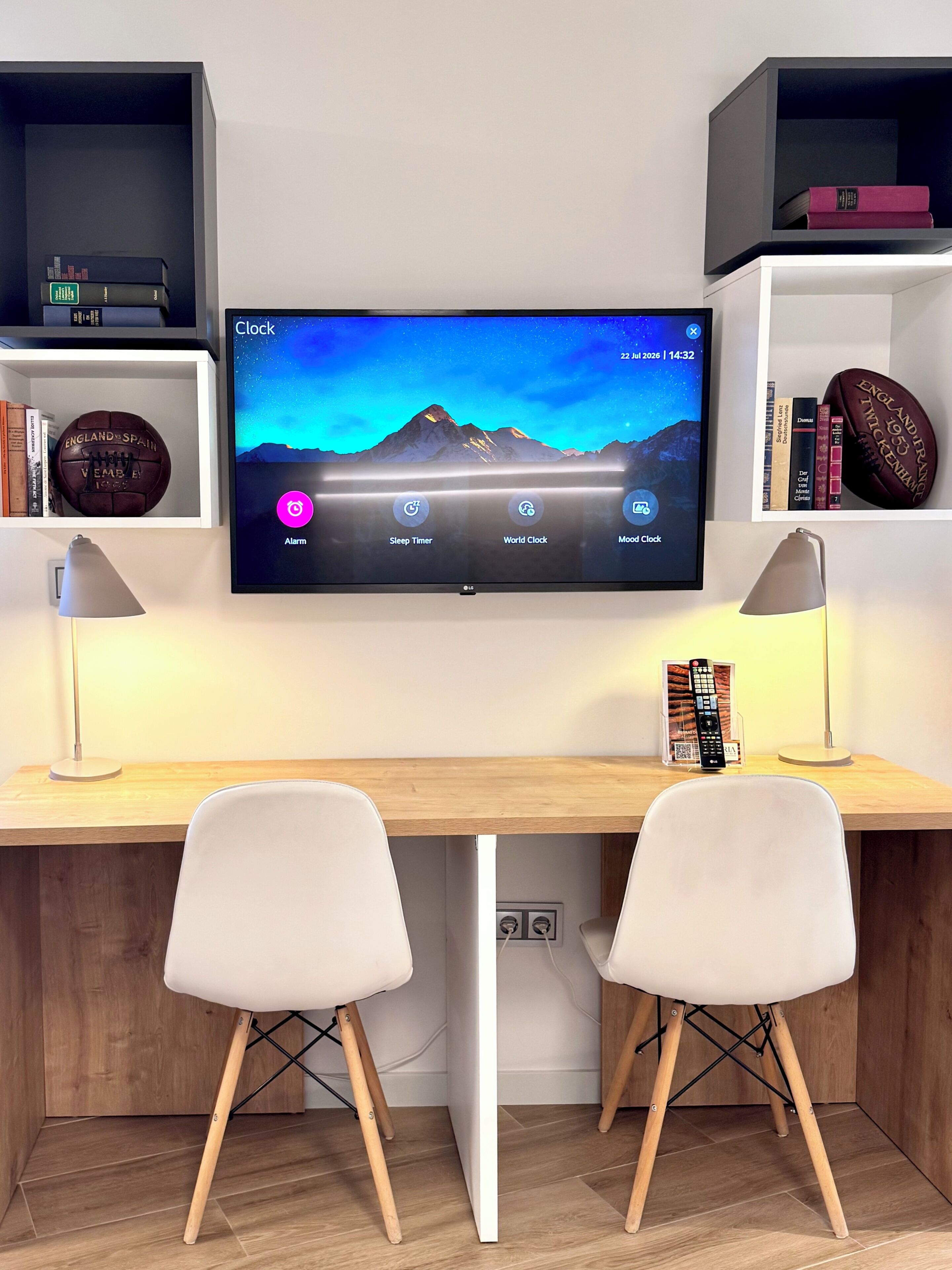 A modern room with a desk, two chairs, a mounted TV displaying a mountain landscape, and shelves with books and decorative items.