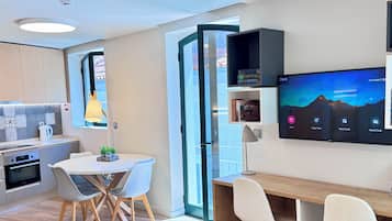 A modern kitchen with a built-in oven, a dining area with a round table and chairs, and a large flat-screen TV mounted on the wall displaying a mountain landscape.