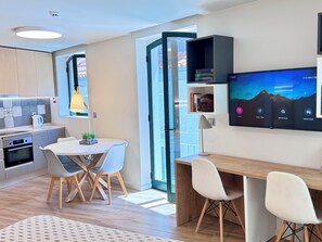 A modern kitchen with a built-in oven, a dining area with a round table and chairs, and a large flat-screen TV mounted on the wall displaying a mountain landscape. - RIA CENTRUM Smart Residence (Aveiro)