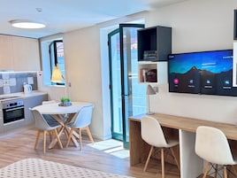 Una cocina moderna con horno integrado, un comedor con mesa redonda y sillas, y un televisor de pantalla plana grande montado en la pared mostrando un paisaje de montaña.