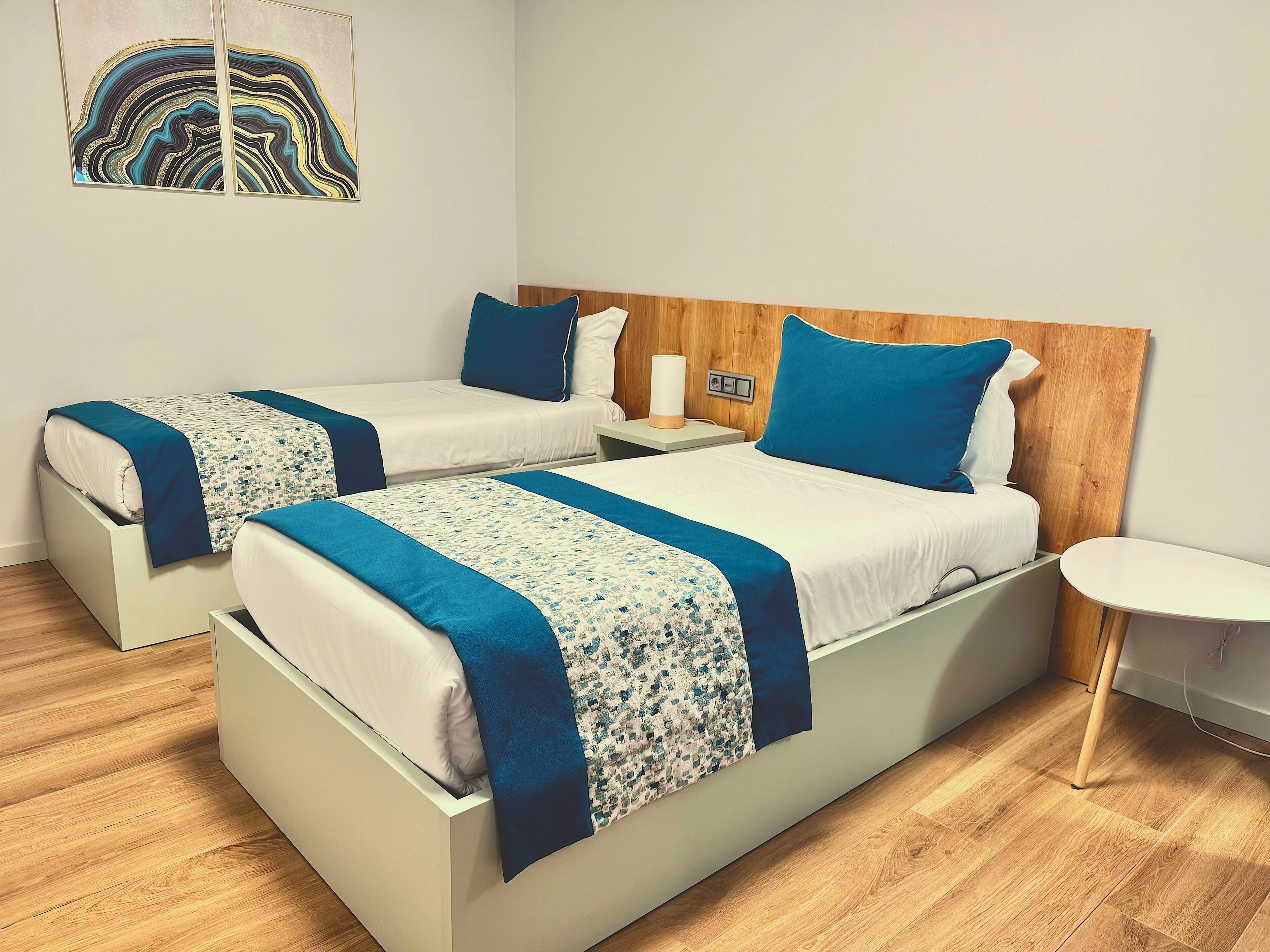 A hotel room with two single beds, a wooden headboard, a bedside table with a lamp, and a small white table.