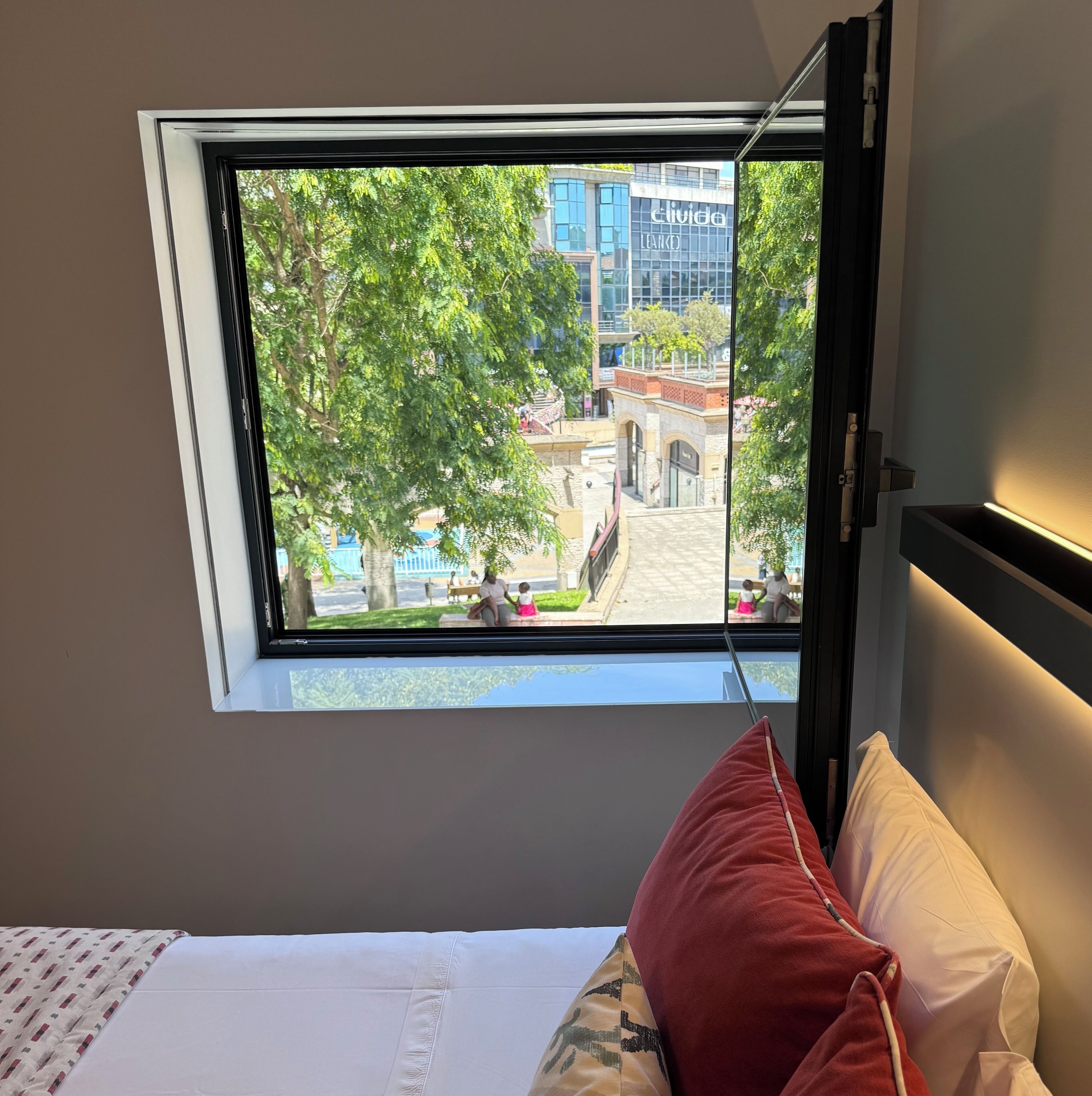 A room with a bed, a window with a view of a building and trees, and a red pillow.