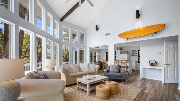 A spacious living area with a large window, a surfboard mounted on the wall, and a cozy seating arrangement. - Wildlife viewers dream! Peaceful & so clean, golf cart incl., pool across street (Fripp Island)