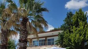 Ein moderner Poolbereich mit Liegestühlen und einer überdachten Lounge, umgeben von Palmen und einem mehrstöckigen Gebäude mit Balkonen.