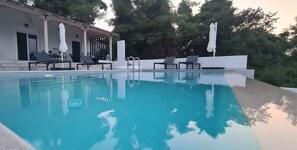 A swimming pool with lounge chairs and umbrellas, surrounded by trees and a building.