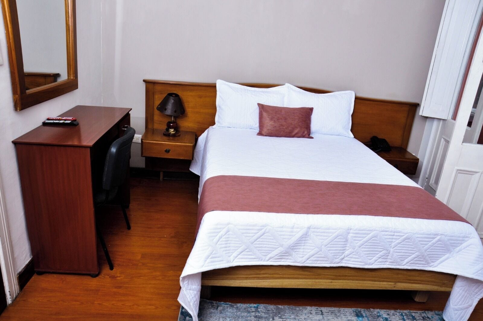 A hotel room with a wooden bed, white bedding, a desk, a chair, a nightstand with a lamp, and a mirror on the wall.