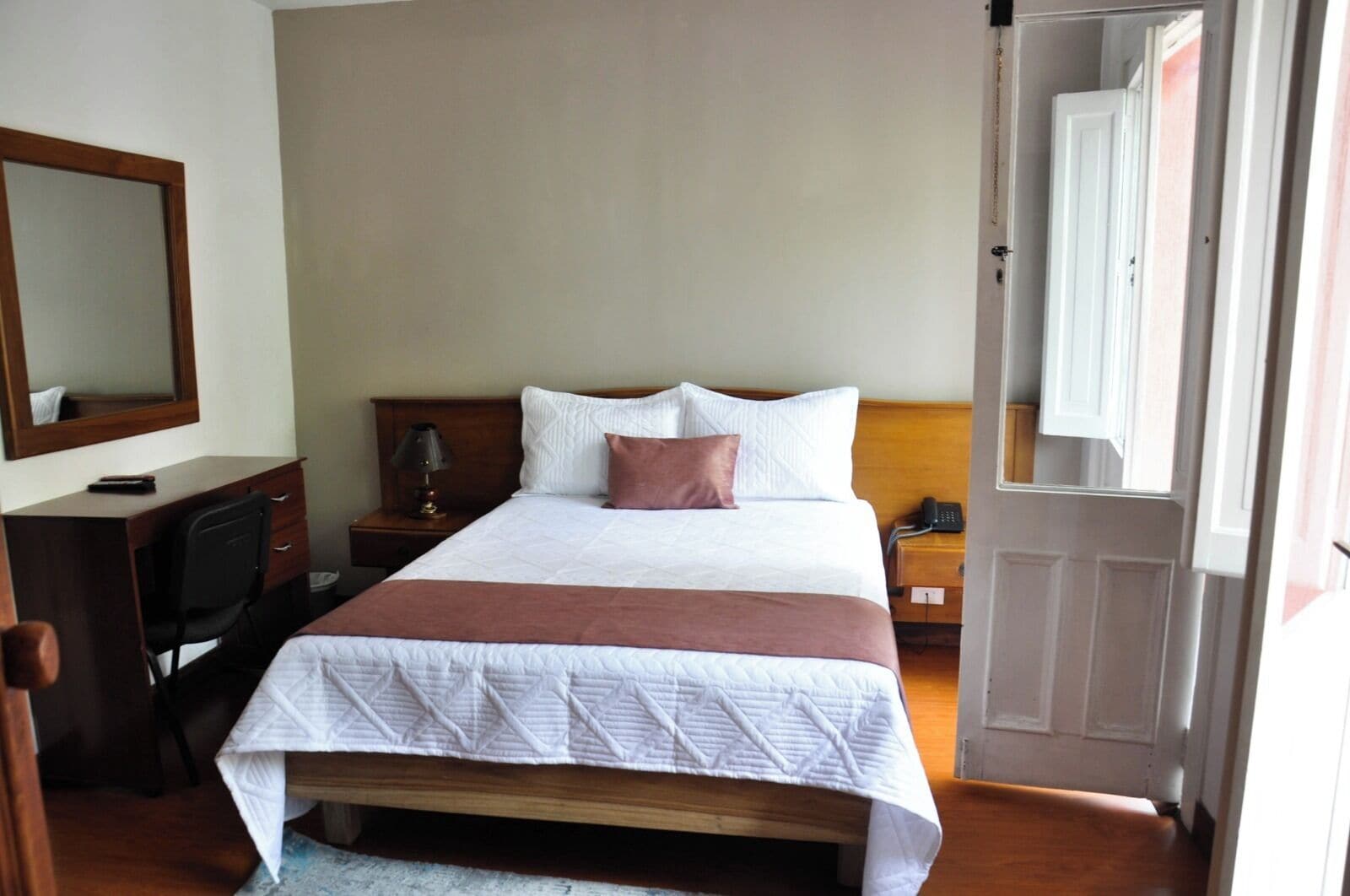 A hotel room with a bed, a desk, a chair, a lamp, and a telephone.