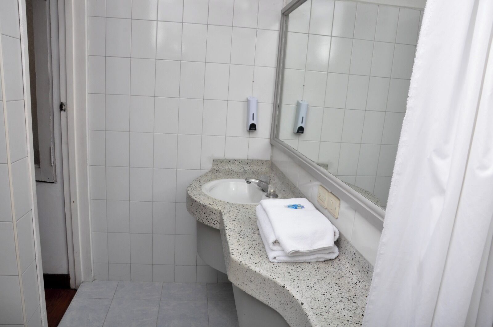 Un baño con paredes de azulejos blancos, encimera de granito, un lavamanos, un espejo y una toalla sobre la encimera.