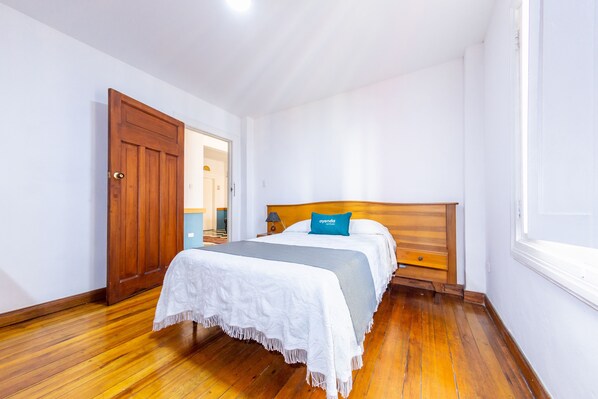 Un dormitorio con piso de madera, cama con ropa de cama blanca y azul, cabecera de madera y una puerta que da a otra habitación.