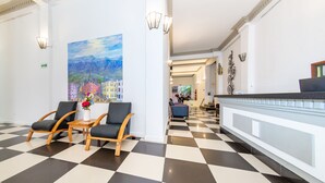 A lobby with a checkered floor, two wooden chairs with black cushions, a small table with a vase of flowers, and a large painting of a cityscape on the wall. - Hotel Escorial (Manizales)