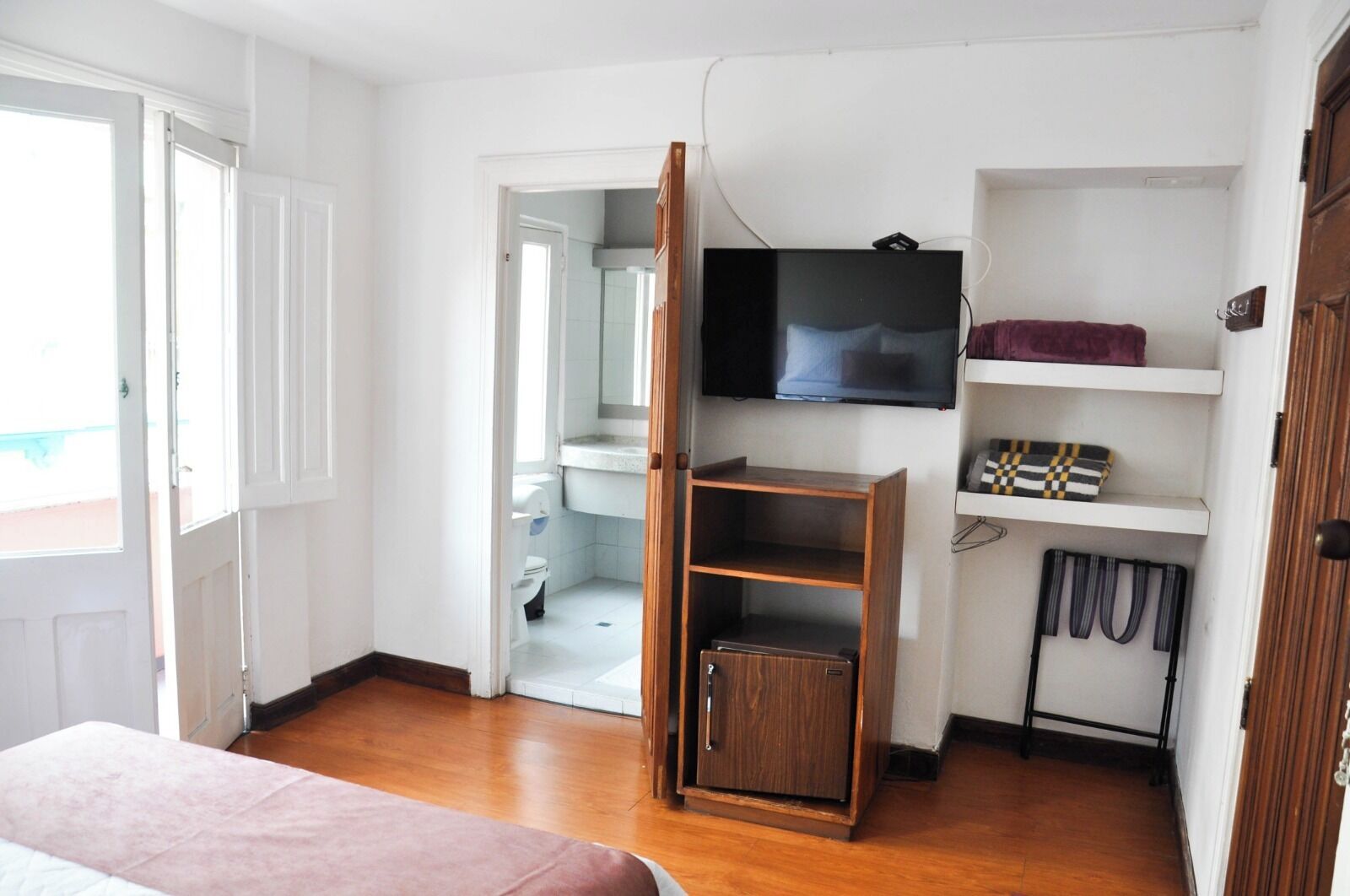 A compact room with a bed, a TV mounted on the wall, a wooden cabinet, and a bathroom visible through an open door.