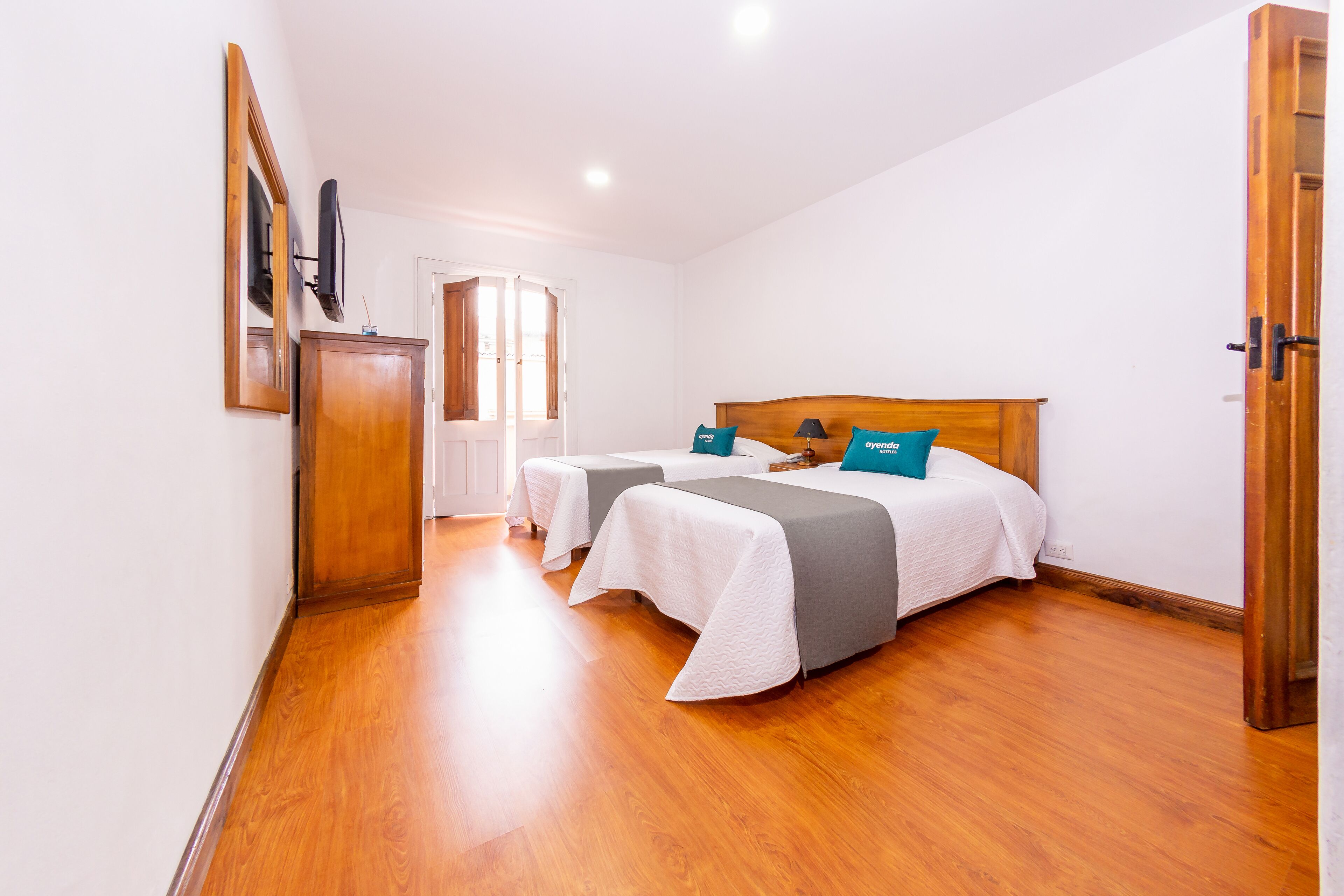 Una habitación de hotel con dos camas, un armario de madera, un televisor montado en la pared y una ventana con cortinas.