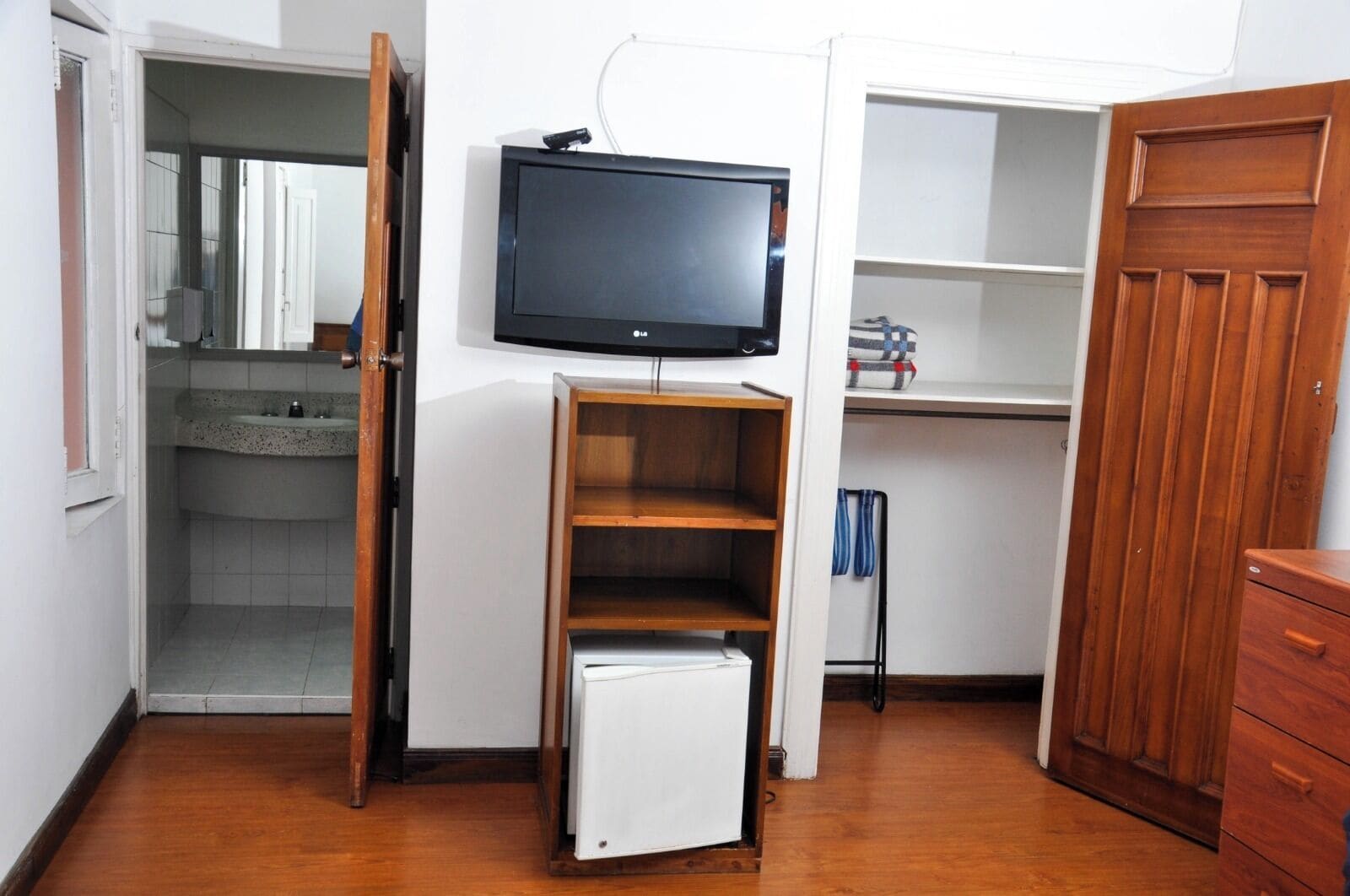 A compact room with a television mounted on the wall, a small refrigerator, and a wooden cabinet.