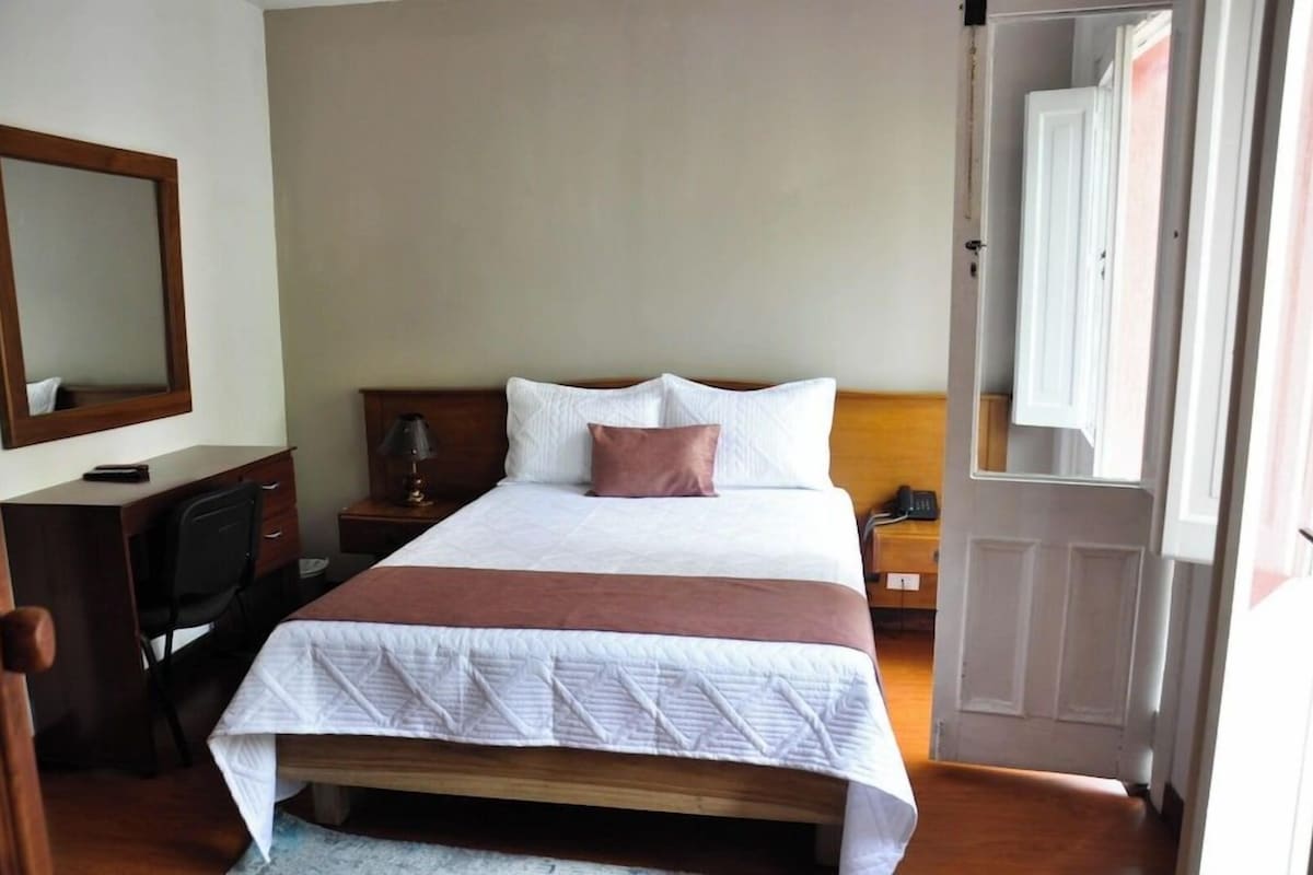 A neatly made bed with a wooden headboard, a bedside table with a lamp, a chair, and a desk with a mirror.