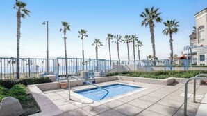 A pool area with a hot tub, surrounded by palm trees and a fence, with a building in the background. - Comfortable, modern condo located steps from the beach! (Oceanside)