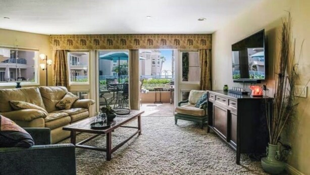 A living room with a sofa, armchair, coffee table, and a TV. There is a balcony with a view of buildings. - Comfortable, modern condo located steps from the beach! (Oceanside)