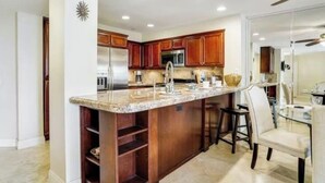 A kitchen with wooden cabinets, a granite countertop, a stainless steel refrigerator, and a dining area with a glass table and chairs. - Comfortable, modern condo located steps from the beach! (Oceanside)