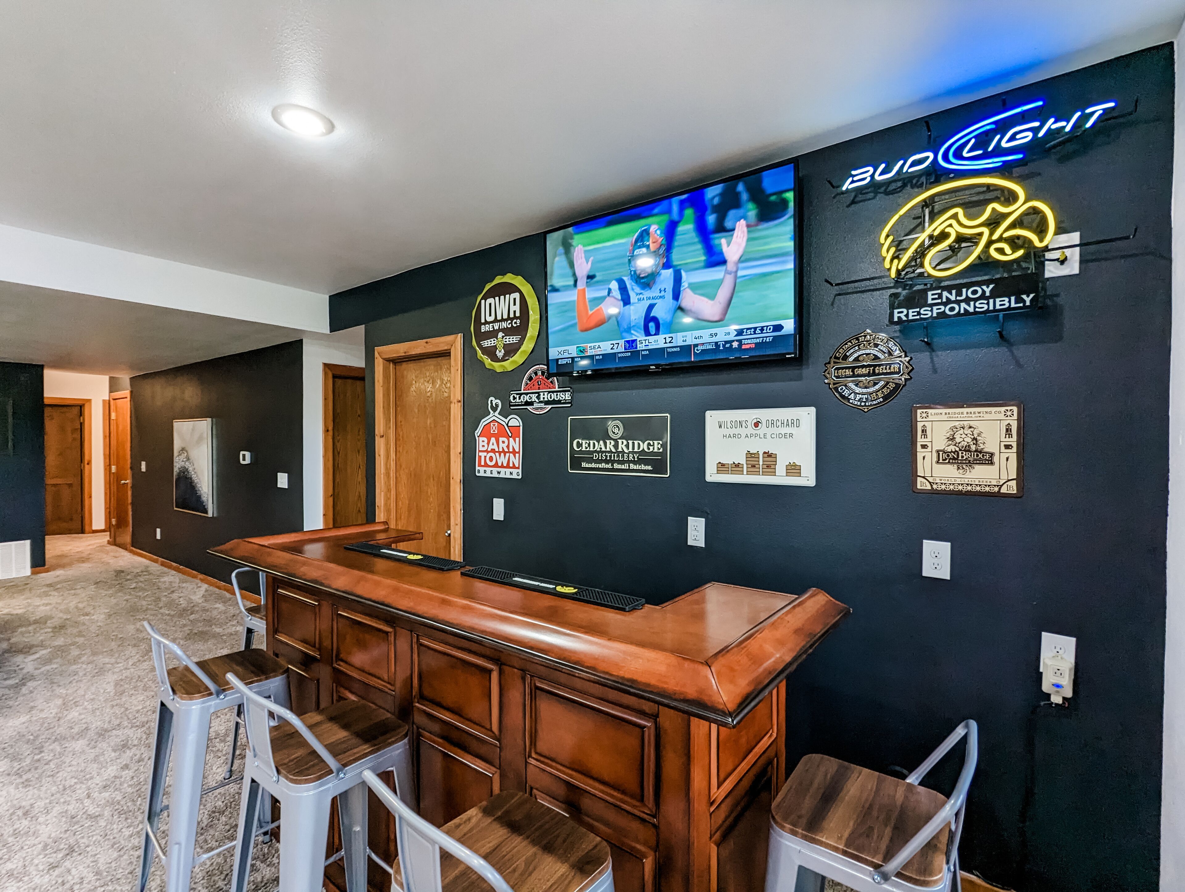 Zona de bar con un televisor mostrando un evento deportivo, rodeada de varios logotipos de cervecerías y destilerías en la pared.