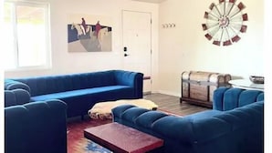 Una sala de estar con un sofá seccional azul, una mesita de centro, un tapete colorido, un ventilador de techo, un reloj y un molino de viento decorativo en la pared.