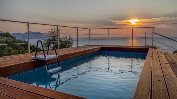 Eine Holzterrasse mit Swimmingpool, einer Leiter und Blick auf den Sonnenuntergang über dem Meer.