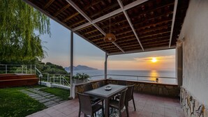 Eine Terrass mit Tisch und Stühlen mit Blick auf eine malerische Landschaft aus Meer und Bergen bei Sonnenuntergang.