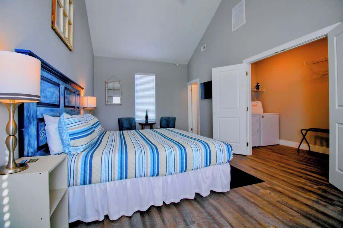 Un dormitorio con cama, mesitas de noche, lámpara, ventana y puerta que da al baño.