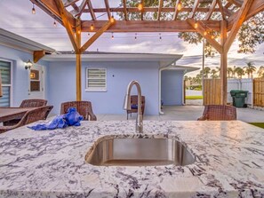 Una cocina con encimera de mármol y pileta de acero inoxidable, bajo un pérgola de madera con luces colgantes.