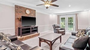 A living room with a fireplace, a television, a ceiling fan, and a large window. - Beautiful Home Walking Distance to Elon University (Elon)