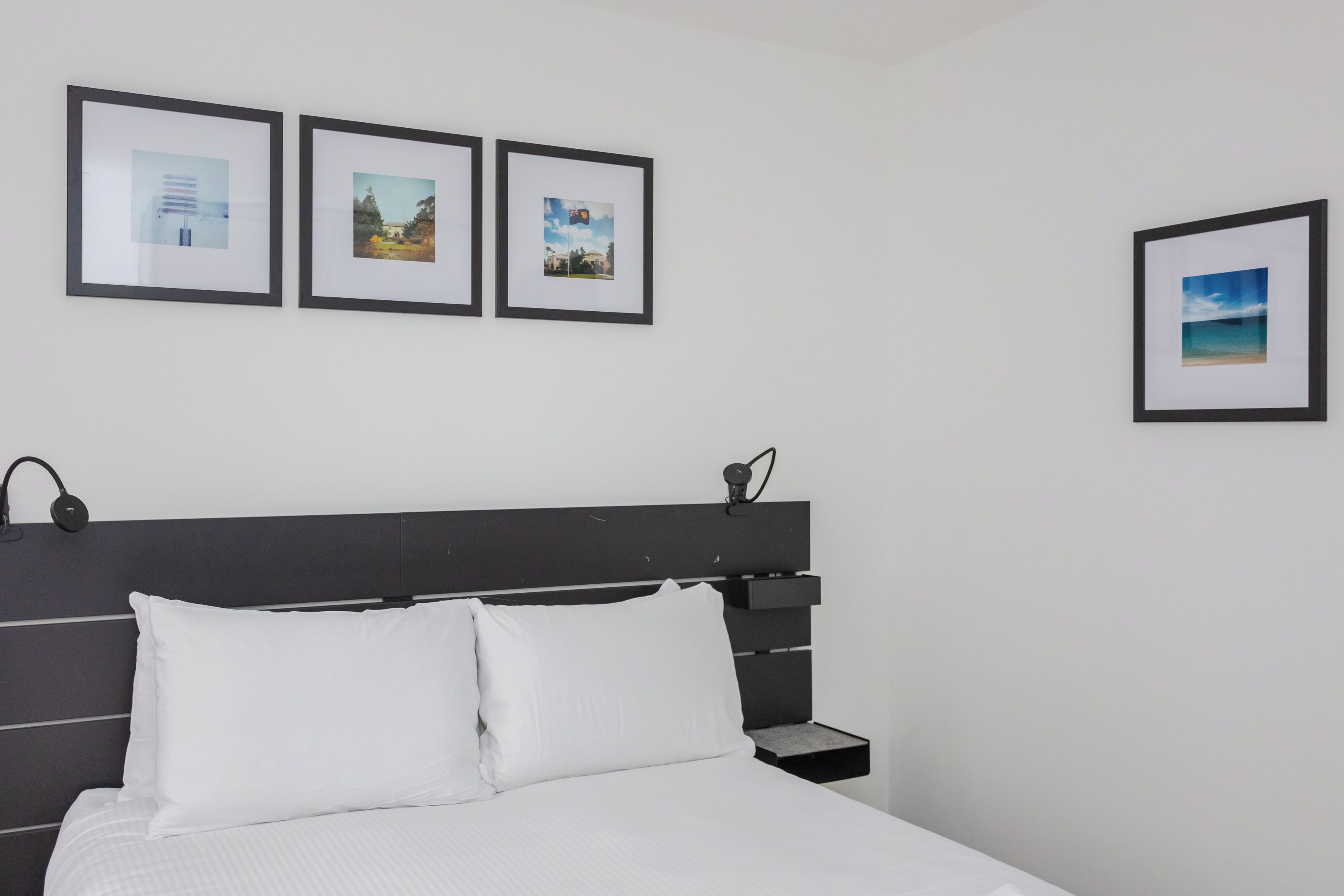 A neatly made bed with white linens, a black headboard, and a bedside table with a lamp. Three framed pictures are hung on the wall above the bed.