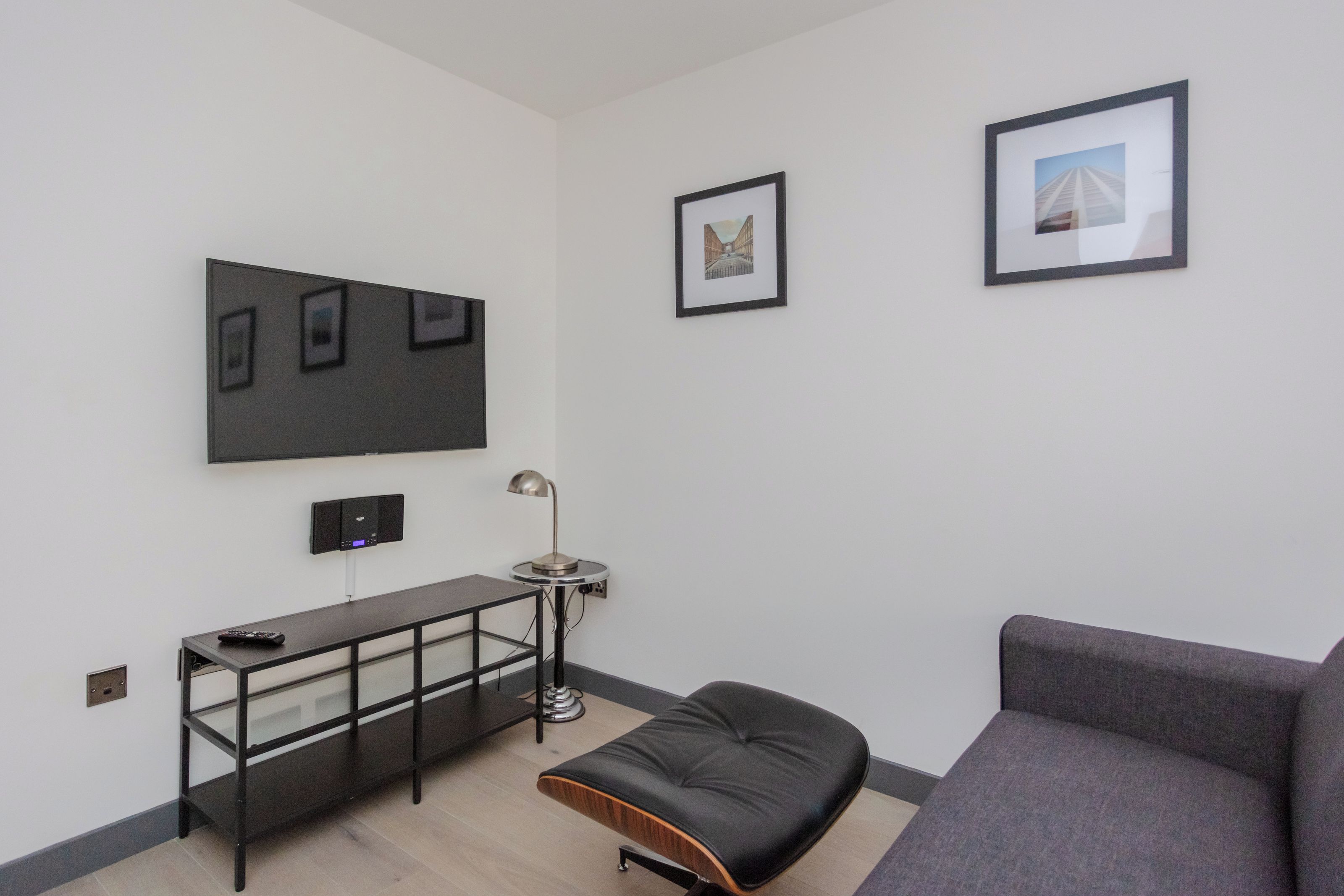 A modern living room with a grey sofa, a black cushioned bench, a small table with a lamp, and a wall-mounted TV.